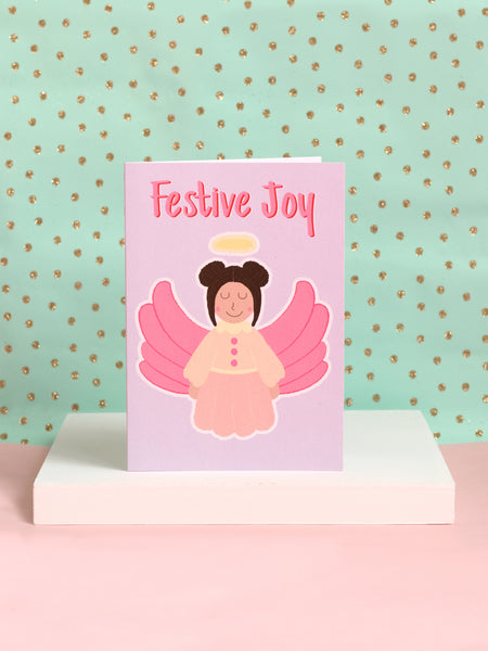 A Christmas card with a pastel dressed angel on the front with the text 'Festive Joy' at the top sat on a white block.